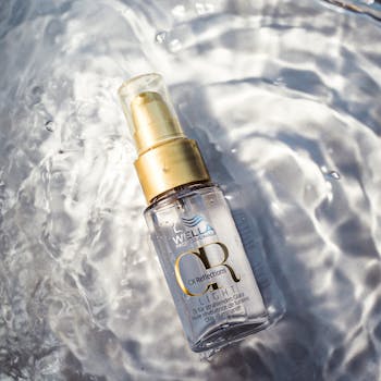 Inicio Transparent hair oil bottle with golden cap on rippling water surface, creating a luxurious reflection effect.